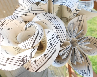 May include: A bouquet of origami flowers made from sheet music. The flowers are white with black musical notes printed on them. The flowers are arranged in a clear glass vase.
