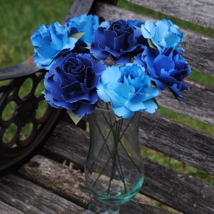 May include: A bouquet of blue and light blue paper roses in a clear glass vase. The roses are arranged in a circular pattern and are sitting on a wooden surface.