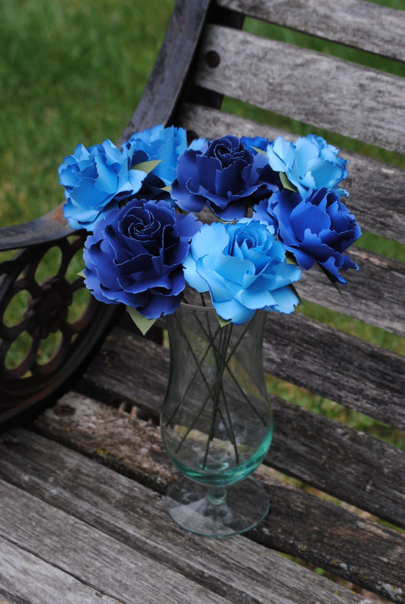 May include: A bouquet of blue and light blue paper roses in a clear glass vase. The roses are arranged in a circular pattern and are sitting on a wooden surface.