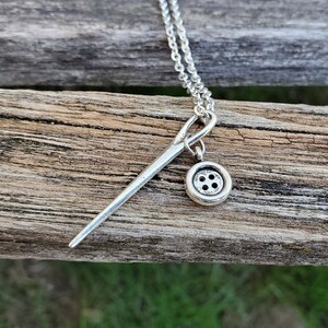 May include: A silver necklace with a needle and button charm. The needle is long and thin, and the button is round with four holes. The necklace is on a wooden surface.