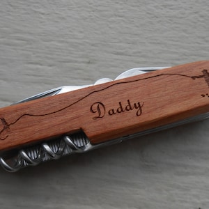 Puede incluir: Un cuchillo de bolsillo de madera con una inscripción personalizada que dice "Daddy". El cuchillo presenta una pequeña imagen grabada de una persona pescando.