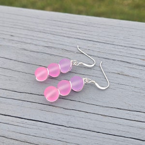 May include: A pair of dangle earrings with three pink and purple glass beads on each earring. The beads are round and have a frosted finish. The earrings are made with silver hooks.