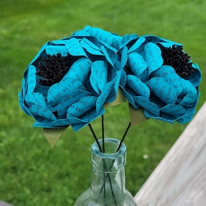 May include: Three teal blue paper flowers with black centers are arranged in a clear glass vase. The flowers are made of paper with handwritten text on them.