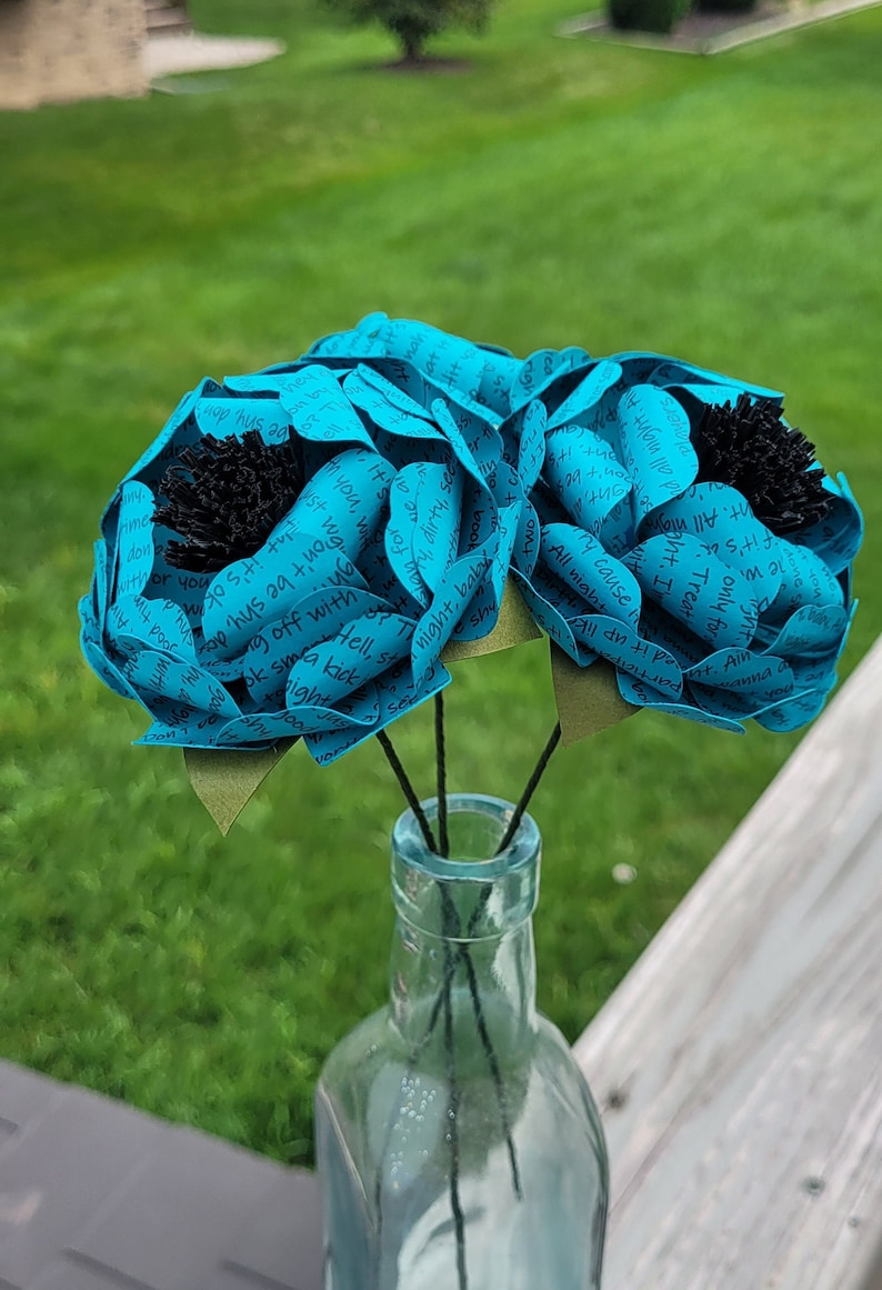 May include: Three teal blue paper flowers with black centers are arranged in a clear glass vase. The flowers are made of paper with handwritten text on them.