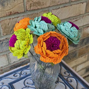 May include: A bouquet of paper flowers in a glass vase. The flowers are made of paper with words printed on them. The flowers are in shades of green, yellow, orange, and pink.