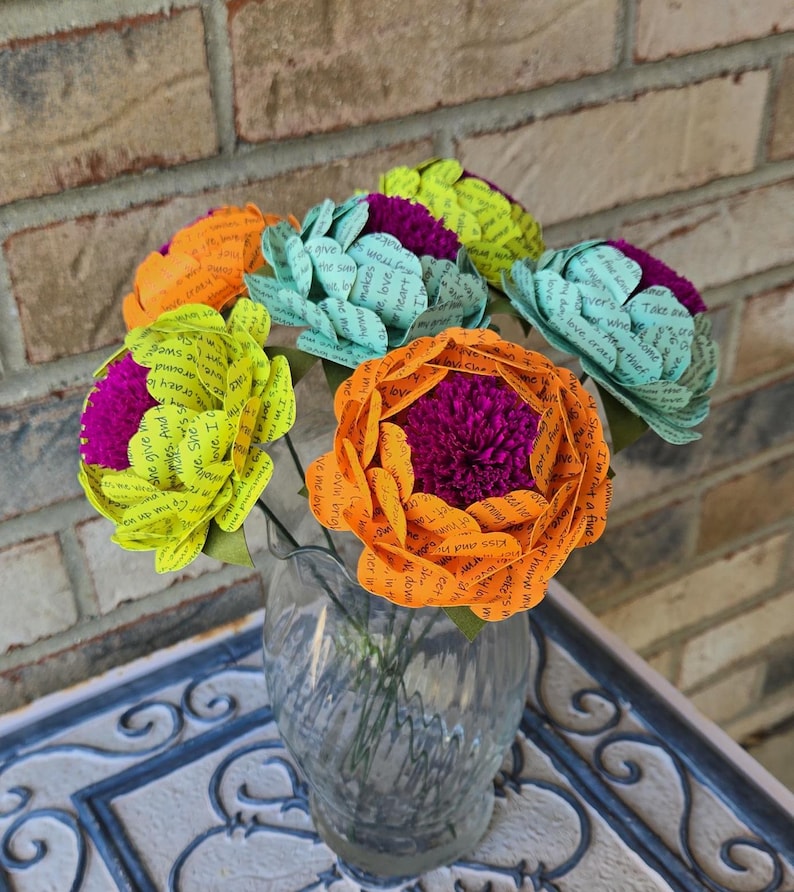 May include: A bouquet of paper flowers in a glass vase. The flowers are made of paper with words printed on them. The flowers are in shades of green, yellow, orange, and pink.