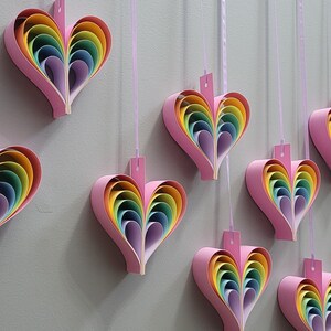 K&ouml;nnte beinhalten: Eine Wand, die mit zehn Regenbogen-Papierherzen geschm&uuml;ckt ist, die an rosa B&auml;ndern h&auml;ngen. Jedes Herz besteht aus &uuml;bereinanderliegenden Papier in den Farben Rot, Orange, Gelb, Gr&uuml;n, Blau, Indigo und Violett.