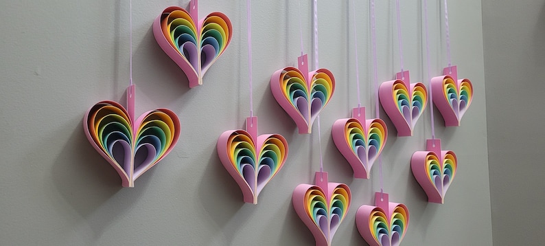 K&ouml;nnte beinhalten: Eine Wand, die mit zehn Regenbogen-Papierherzen geschm&uuml;ckt ist, die an rosa B&auml;ndern h&auml;ngen. Jedes Herz besteht aus &uuml;bereinanderliegenden Papier in den Farben Rot, Orange, Gelb, Gr&uuml;n, Blau, Indigo und Violett.