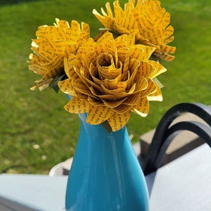 May include: Three yellow paper flowers with black text printed on them are arranged in a blue vase. The text on the flowers includes phrases like "Til you leave the sight of me", "I'll never form", and "Like a stately cloak".