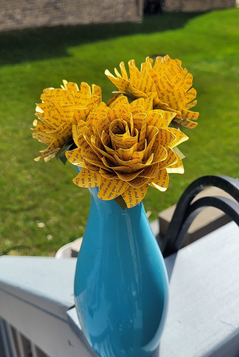 May include: Three yellow paper flowers with black text printed on them are arranged in a blue vase. The text on the flowers includes phrases like "Til you leave the sight of me", "I'll never form", and "Like a stately cloak".