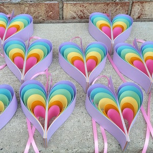 K&ouml;nnte beinhalten: Neun regenbogenfarbene, mehrschichtige Papierherzen mit einem rosa Band an jedem. Die Herzen bestehen aus mehreren Papierschichten in den Farben Rosa, Orange, Gelb, Gr&uuml;n und Blau.