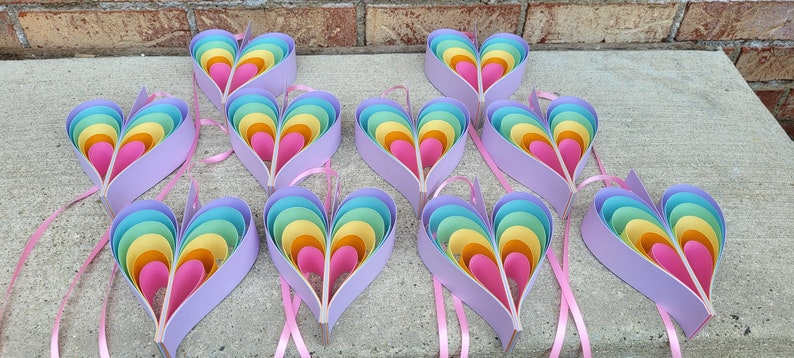 K&ouml;nnte beinhalten: Neun regenbogenfarbene, mehrschichtige Papierherzen mit einem rosa Band an jedem. Die Herzen bestehen aus mehreren Papierschichten in den Farben Rosa, Orange, Gelb, Gr&uuml;n und Blau.