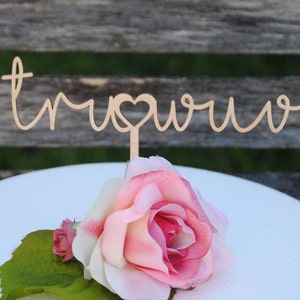 May include: A wooden cake topper with the words "true love" in cursive script with a heart in the middle of the word "love". The topper is on a white cake with a pink rose.