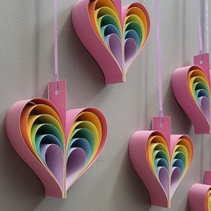 K&ouml;nnte beinhalten: F&uuml;nf rosa Papierherzen mit Regenbogen-Schichten, die an einer wei&szlig;en Schnur h&auml;ngen. Jedes Herz besteht aus mehreren Schichten Papier in Regenbogenfarben, von Lila bis Rot.