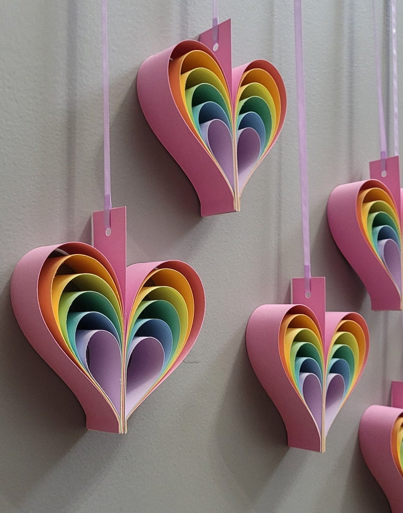 K&ouml;nnte beinhalten: F&uuml;nf rosa Papierherzen mit Regenbogen-Schichten, die an einer wei&szlig;en Schnur h&auml;ngen. Jedes Herz besteht aus mehreren Schichten Papier in Regenbogenfarben, von Lila bis Rot.