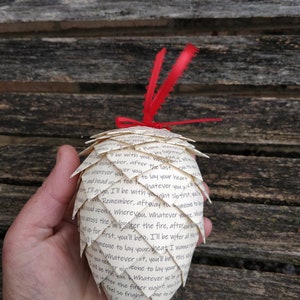 Pode incluir: Um enfeite de papel branco em forma de pinha, decorado com uma fita vermelha e uma pena. O enfeite &eacute; feito de p&aacute;ginas de um livro e apresenta um texto escrito &agrave; m&atilde;o que diz: "Consomme flame. Viator by light I'll be with you, who bsingimber, after the last. ver you er wherever you tand head up too fo to lay your heart Whatever you tr. Ill gingo, I'll be with youight slo tirst, youle Dyrevo Remember, aft way to homeone to la Ирои, wherevou. Whatever youge. Wh wcross the I'm lones I'm pinter the fire, after mot up for first, you'll beo, I'll be witer all ther bomeone to lay your heat I wa You whatever ist, you'll be this vid I will be the you. Whatever year creone to lay your tiv ve itfter the fireer niglet your feel so frigher one to lay St. Can't Jive it what will be teve"