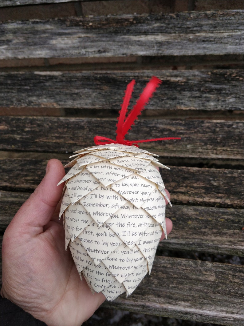 Pode incluir: Um enfeite de papel branco em forma de pinha, decorado com uma fita vermelha e uma pena. O enfeite &eacute; feito de p&aacute;ginas de um livro e apresenta um texto escrito &agrave; m&atilde;o que diz: "Consomme flame. Viator by light I'll be with you, who bsingimber, after the last. ver you er wherever you tand head up too fo to lay your heart Whatever you tr. Ill gingo, I'll be with youight slo tirst, youle Dyrevo Remember, aft way to homeone to la Ирои, wherevou. Whatever youge. Wh wcross the I'm lones I'm pinter the fire, after mot up for first, you'll beo, I'll be witer all ther bomeone to lay your heat I wa You whatever ist, you'll be this vid I will be the you. Whatever year creone to lay your tiv ve itfter the fireer niglet your feel so frigher one to lay St. Can't Jive it what will be teve"