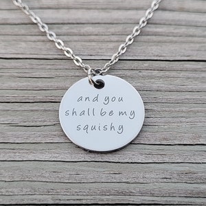 May include: A silver necklace featuring a circular pendant inscribed with the words "and you shall be my squishy". The chain is a delicate link design. The pendant is made of a shiny metal.