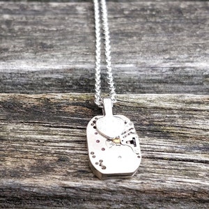 May include: A silver necklace with a pendant featuring a heart-shaped design and a clockwork mechanism. The pendant is hanging on a wooden surface.