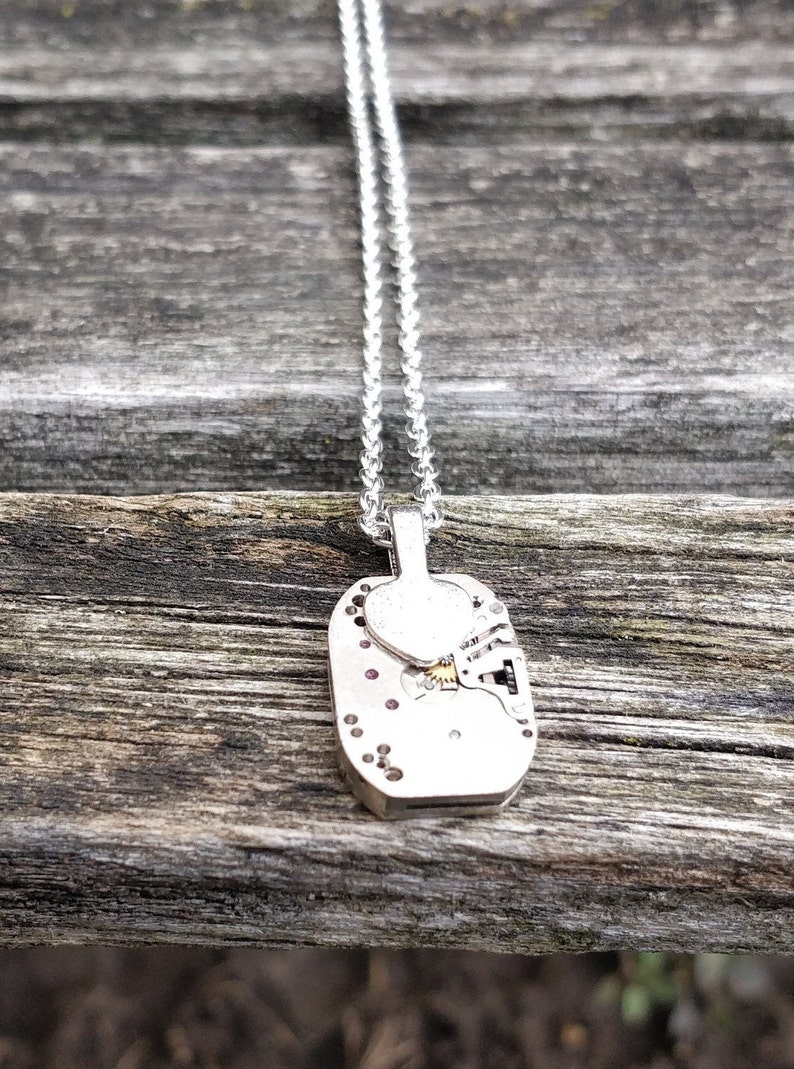 May include: A silver necklace with a pendant featuring a heart-shaped design and a clockwork mechanism. The pendant is hanging on a wooden surface.