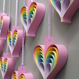 K&ouml;nnte beinhalten: Ein Set aus sieben rosa Papierherzen mit regenbogenfarbenen, &uuml;bereinanderliegenden Papierbl&auml;ttern im Inneren. Die Herzen h&auml;ngen an einem wei&szlig;en Band.