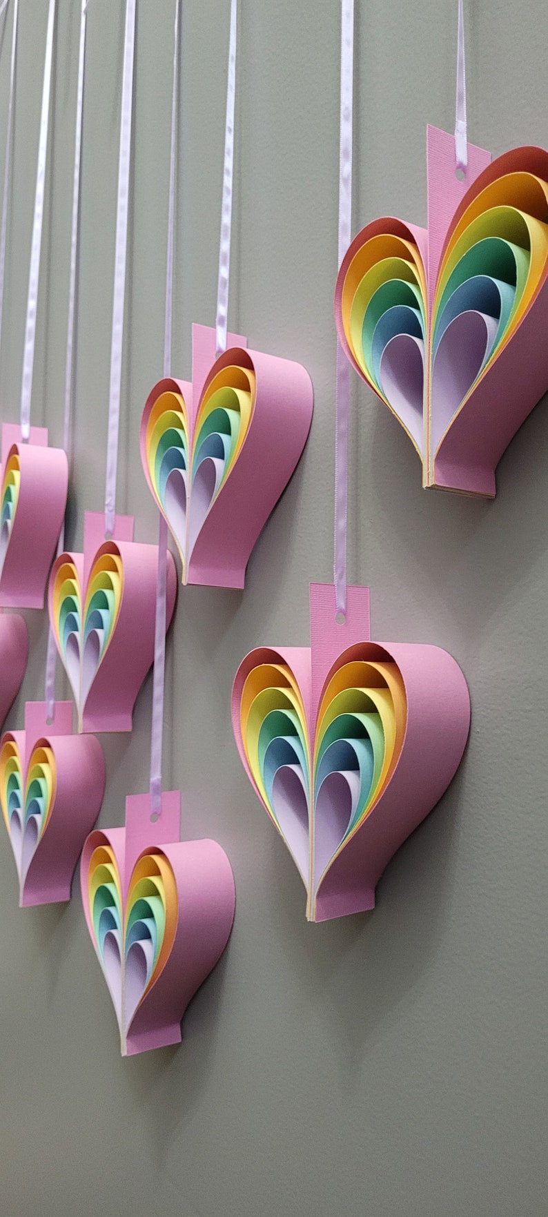 K&ouml;nnte beinhalten: Ein Set aus sieben rosa Papierherzen mit regenbogenfarbenen, &uuml;bereinanderliegenden Papierbl&auml;ttern im Inneren. Die Herzen h&auml;ngen an einem wei&szlig;en Band.