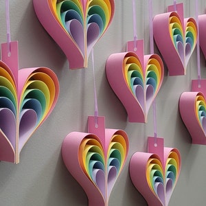 K&ouml;nnte beinhalten: Ein Set aus acht rosa Papierherzen mit regenbogenfarbenen, &uuml;bereinanderliegenden Papierbl&auml;ttern im Inneren. Die Herzen h&auml;ngen an einer wei&szlig;en Wand mit einer Schnur.