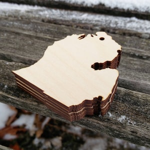 May include: A stack of wooden cutouts in the shape of the state of Michigan. The cutouts are made of light-colored wood with dark brown edges. The stack is resting on a weathered wooden surface, with snow in the background.