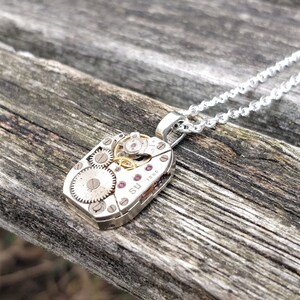 May include: A silver necklace with a pendant made from a vintage watch face. The pendant features exposed gears and a small ruby-colored stone. The necklace is on a wooden surface.