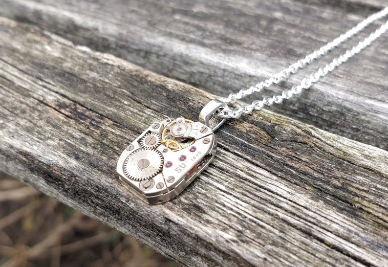 May include: A silver necklace with a pendant made from a vintage watch face. The pendant features exposed gears and a small ruby-colored stone. The necklace is on a wooden surface.