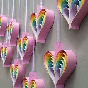 K&ouml;nnte beinhalten: Eine Reihe von regenbogenf&ouml;rmigen Herz-Papierornamenten, die an einem wei&szlig;en Band h&auml;ngen. Jedes Herz besteht aus &uuml;bereinanderliegendem Papier in den Farben Rosa, Gelb, Gr&uuml;n, Blau und Orange.