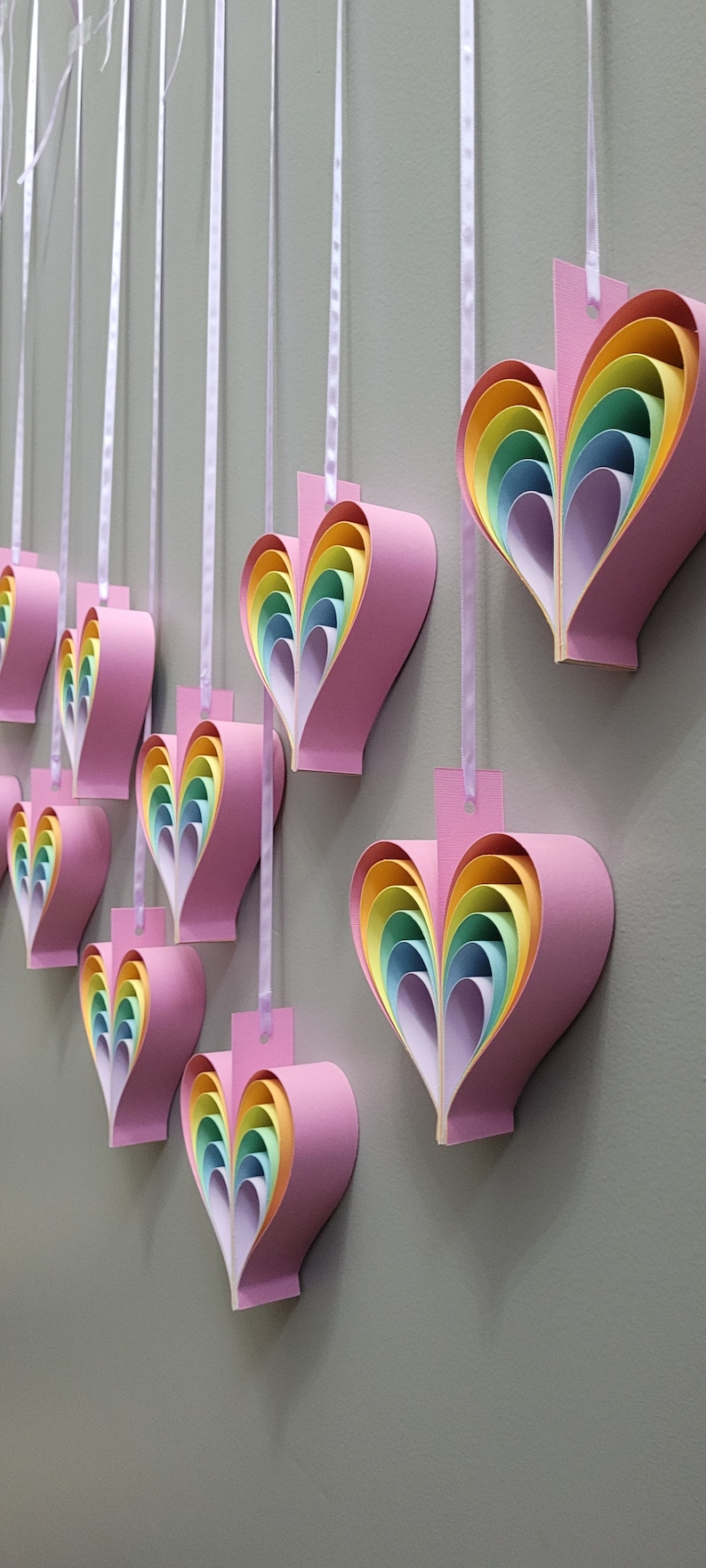 K&ouml;nnte beinhalten: Eine Reihe von regenbogenf&ouml;rmigen Herz-Papierornamenten, die an einem wei&szlig;en Band h&auml;ngen. Jedes Herz besteht aus &uuml;bereinanderliegendem Papier in den Farben Rosa, Gelb, Gr&uuml;n, Blau und Orange.