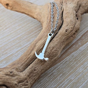 May include: A silver hammer charm necklace with a silver chain. The necklace is displayed on a piece of driftwood.