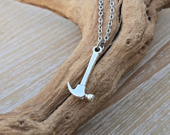 Op de afbeelding: Een zilveren hamer hanger ketting met een zilveren ketting. De ketting ligt op een stuk drijfhout.