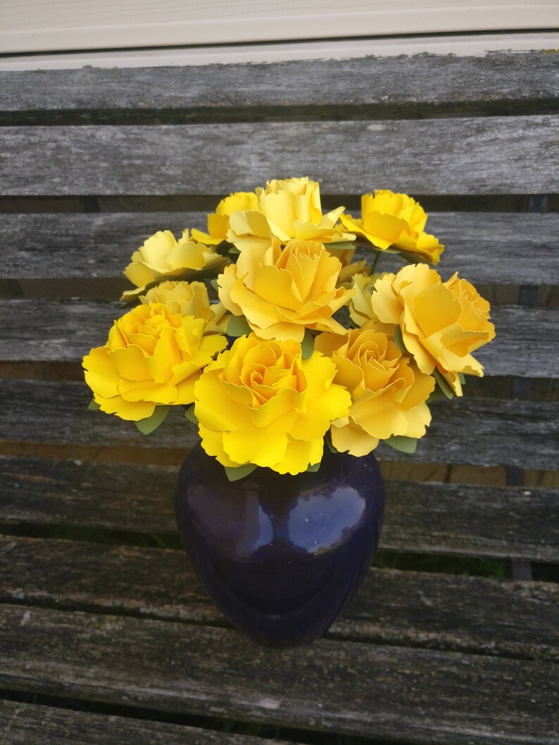 May include: A bouquet of yellow paper flowers in a dark purple vase. The vase is sitting on a wooden surface.