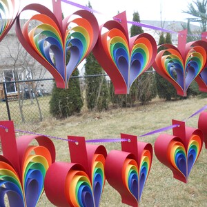 May include: A string of rainbow-colored paper hearts hanging from a purple ribbon. The hearts are made of multiple layers of paper, creating a 3D effect. The hearts are hanging in front of a green lawn and a fence.