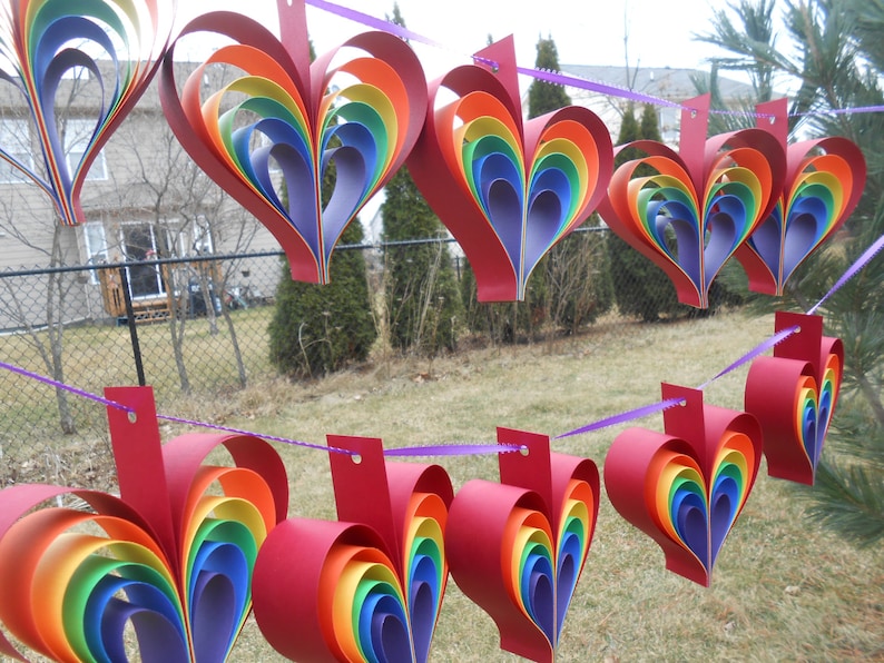 May include: A string of rainbow-colored paper hearts hanging from a purple ribbon. The hearts are made of multiple layers of paper, creating a 3D effect. The hearts are hanging in front of a green lawn and a fence.