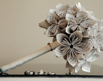 May include: A bouquet of white paper flowers made from the pages of a book. The flowers are arranged in a circular shape and are held together by a white ribbon. The bouquet is sitting on a black surface with three silver rings.