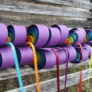 May include: A rainbow paper heart garland with purple paper hearts and colorful ribbon streamers. The garland is hanging on a wooden bench.
