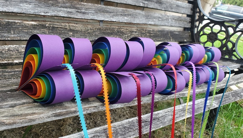 May include: A rainbow paper heart garland with purple paper hearts and colorful ribbon streamers. The garland is hanging on a wooden bench.