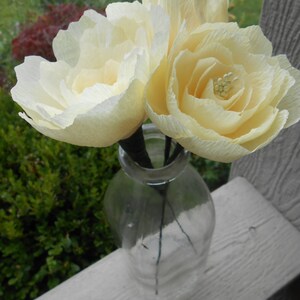 May include: Two cream-colored paper flowers in a clear glass bottle with a textured surface.