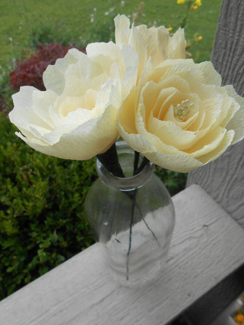 May include: Two cream-colored paper flowers in a clear glass bottle with a textured surface.