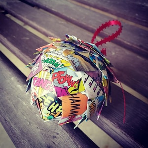 May include: A colorful, handmade ornament made from comic book pages. The ornament is shaped like a sphere and features various comic book words and phrases, including "POW!" and "BAM!" The ornament is decorated with a red ribbon.