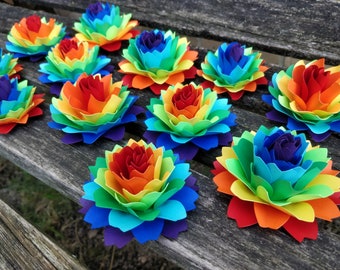 May include: A collection of rainbow paper flowers arranged on a wooden surface. Each flower is made of multiple layers of paper in shades of red, orange, yellow, green, blue, indigo, and violet.