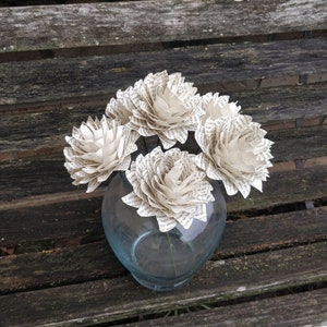 May include: Four white paper flowers made from book pages are arranged in a clear glass vase. The vase is sitting on a wooden bench.