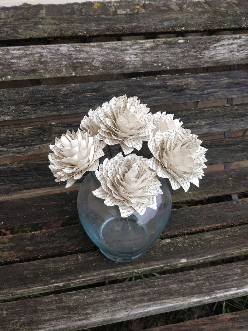 May include: Four white paper flowers made from book pages are arranged in a clear glass vase. The vase is sitting on a wooden bench.