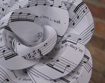 May include: A white paper rose made from sheet music. The rose is made of several layers of paper, each with a different section of music printed on it. The text on the paper includes the lyrics "I can see your", "Well, ba-by, they're", "hing I need", "for-get", "ly nev", "his don't e-".