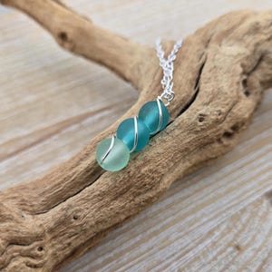 Puede incluir: Un collar con tres cuentas de vidrio marino en tonos turquesa y verde, envueltas con alambre de plata. Las cuentas están ensartadas en una delicada cadena de plata. El collar se exhibe sobre un trozo de madera flotante.