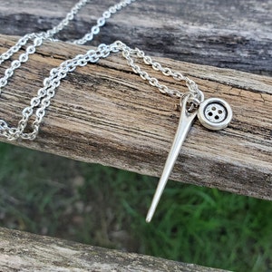 May include: A silver necklace with a needle and button charm. The needle is long and thin, and the button is round with four holes. The chain is a simple silver chain.