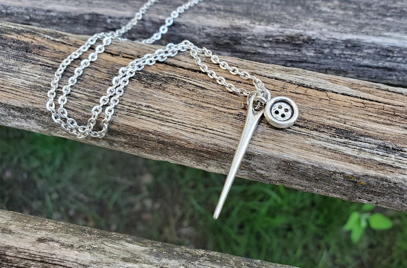 May include: A silver necklace with a needle and button charm. The needle is long and thin, and the button is round with four holes. The chain is a simple silver chain.